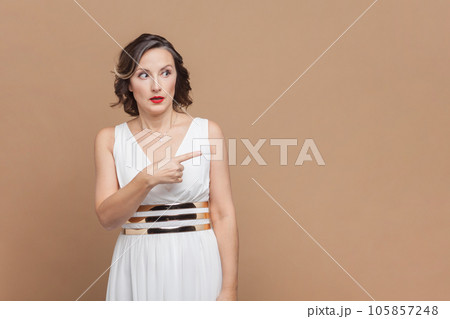 Portrait of shocked surprised middle aged woman with wavy hair pointing away at copy space for advertisement, wearing white dress. Indoor studio shot isolated on light brown background. 105857248