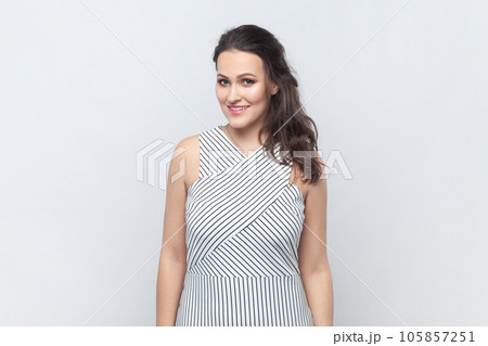 Portrait of attractive adorable smiling brunette woman standing looking at camera, being in good mood, expressing happiness. wearing striped dress. Indoor studio shot isolated on gray background. 105857251