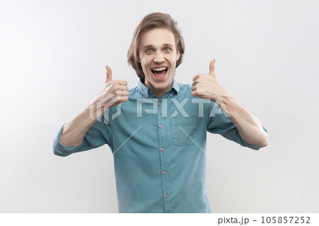 Portrait of excited amazed optimistic cheerful young man showing thumbs up, screaming with happiness, recommend service, wearing blue shirt. Indoor studio shot isolated on gray background. 105857252