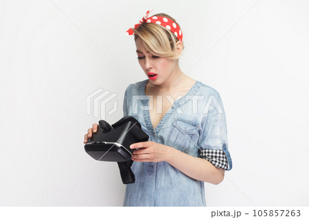 Portrait of concentrated blonde woman wearing blue denim shirt and red headband standing holding vr glasses, equipment for gaming. Indoor studio shot isolated on gray background. Portrait of concentrated blonde woman wearing blue denim shirt and red headband standing holding vr glasses, equipment for gaming. Indoor studio shot isolated on gray background. 105857263
