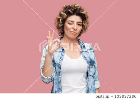 Portrait of playful woman with curly hairstyle wearing blue shirt standing showing okay gesture, winking to camera, saying great job. Indoor studio shot isolated on pink background. 105857286