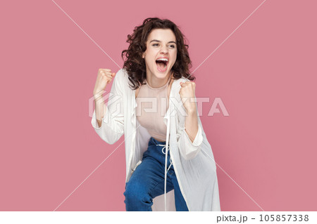 Portrait of excited overjoyed young adult woman with curly hair wearing casual style outfit clenched her fists celebrating victory, expressing happiness. Indoor studio shot isolated on pink background 105857338