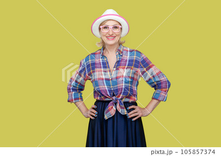 Cheerful positive optimistic senior woman wearing checkered shirt, hat and eyeglasses standing with hands on hips, looking at camera with happiness. Indoor studio shot isolated on yellow background. Cheerful positive optimistic senior woman wearing checkered shirt, hat and eyeglasses standing with hands on hips, looking at camera with happiness. Indoor studio shot isolated on yellow background. 105857374