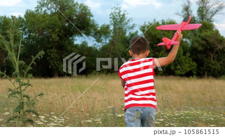 Brunet boy plays with toy plane dreaming to be pilot on green meadow backside view. Happy little boy with toy plane runs across sunset field. Boy with toy enjoys summer vacation at evening twilight 105861515