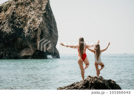 Woman and her daughter practicing balancing yoga pose on one leg up together on rock in the sea. Silhouette mother and daughter doing yoga at beach Woman and her daughter practicing balancing yoga pose on one leg up together on rock in the sea. Silhouette mother and daughter doing yoga at beach 105866048