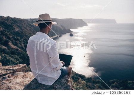 Digital nomad, man in the hat, a businessman with a laptop sits on the rocks by the sea during sunset, makes a business transaction online from a distance. Remote work on vacation. Digital nomad, man in the hat, a businessman with a laptop sits on the rocks by the sea during sunset, makes a business transaction online from a distance. Remote work on vacation. 105866168