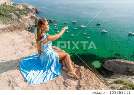 Woman travel sea. Happy woman in a beautiful location poses on a cliff high above the sea, with emerald waters and yachts in the background, while sharing her travel experiences Woman travel sea. Happy woman in a beautiful location poses on a cliff high above the sea, with emerald waters and yachts in the background, while sharing her travel experiences 105868211