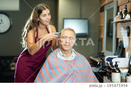 Young woman hairdresser performs haircut for elderly man using hair clipper Young woman hairdresser performs haircut for elderly man using hair clipper 105875956