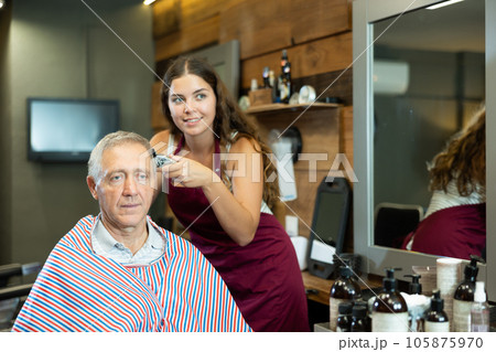 Young woman hairdresser performs haircut for elderly man using hair clipper Young woman hairdresser performs haircut for elderly man using hair clipper 105875970