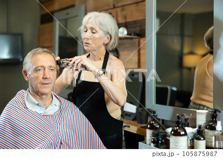 Haircut in a stylish barbershop - an elderly man sits in a chair, woman cuts her hair and does hair styling Haircut in a stylish barbershop - an elderly man sits in a chair, woman cuts her hair and does hair styling 105875987