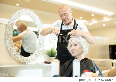 Experienced senior man master makes performs haircut for mature woman with scissors Experienced senior man master makes performs haircut for mature woman with scissors 105876607