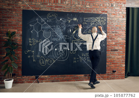 Young bald man, scientist, professor standing by blackboard with chemical formulas and calculations. Making research Young bald man, scientist, professor standing by blackboard with chemical formulas and calculations. Making research 105877762