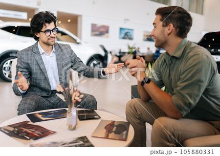 Happy male customer talking about terms of buying car with manager wearing business suit sitting at desk in auto dealership. Concept of choosing, buying new auto at showroom. 105880331