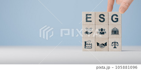 ESG concept of environmental, social and governance. Hand holds wooden cubes with abbreviation ESG standing with other ESG icons on blue background. Copy space. wide banner 105881096