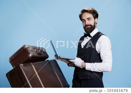Doorman with laptop in hand showing expression of hotel industry, showcases proficiency in utilizing technology to offer assistance. Bellhop in suit making reservations or provides information. 105881679