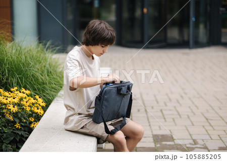 Boy open laptop bag and takes out his laptop. Boy sits on bench outdoors 105885205