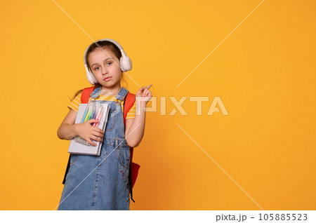 Smart first grade student girl in headphones and casual denim overalls, points aside a copy advertising space for text Smart first grade student girl in headphones and casual denim overalls, points aside a copy advertising space for text 105885523