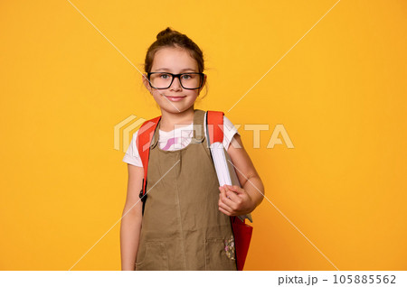 Smiling school girl carrying a backpack and textbook, expressing positive emotions, happy and pride to be first grader 105885562