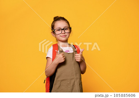 Adorable little kid girl in casual clothes and glasses, carrying her backpack, gestures with thumbs up looking at camera Adorable little kid girl in casual clothes and glasses, carrying her backpack, gestures with thumbs up looking at camera 105885606