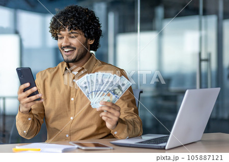 Businessman at workplace happy with financial achievement results, holding phone and dollar bill money in hands, man working sitting with laptop inside office. Businessman at workplace happy with financial achievement results, holding phone and dollar bill money in hands, man working sitting with laptop inside office. 105887121