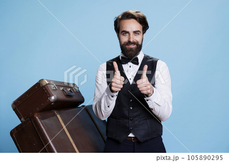 Smiling bellboy does thumbs up sign while he is wearing formal hotel attire in studio, expressing success and agreement. Young man dressed as doorman shows like symbol, tourism industry. Smiling bellboy does thumbs up sign while he is wearing formal hotel attire in studio, expressing success and agreement. Young man dressed as doorman shows like symbol, tourism industry. 105890295