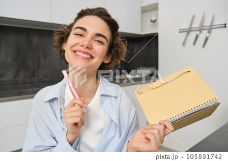 Portrait of smiling brunette girl, holds her planner, writes in notebook and looks happy, laughs, does homework, makes notes, sits at home in kitchen Portrait of smiling brunette girl, holds her planner, writes in notebook and looks happy, laughs, does homework, makes notes, sits at home in kitchen 105891742