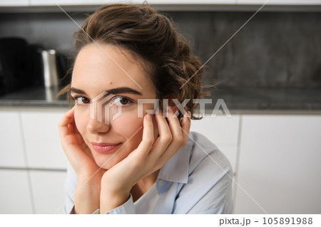 Close up portrait of young woman, 25 years old, sitting in her kitchen alone, express candid happiness, smiles, leans head on hands Close up portrait of young woman, 25 years old, sitting in her kitchen alone, express candid happiness, smiles, leans head on hands 105891988