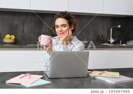 Self-employed woman works from homes, sits with laptop and drinks coffee in kitchen, surrounded with documents. Girl student studies remote, does project on computer, holds a cup Self-employed woman works from homes, sits with laptop and drinks coffee in kitchen, surrounded with documents. Girl student studies remote, does project on computer, holds a cup 105891989