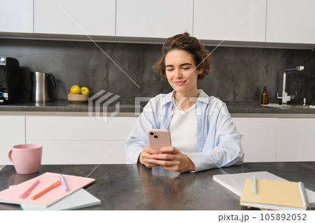 Technology and lifestyle. Young woman sits at home, uses smartphone in her kitchen and smiles 105892052