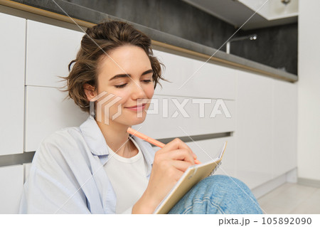 Close up portrait of young woman writing in her journal, feeling creative, sitting with planner, notebook and pen on floor, studying, doing her homework at home Close up portrait of young woman writing in her journal, feeling creative, sitting with planner, notebook and pen on floor, studying, doing her homework at home 105892090