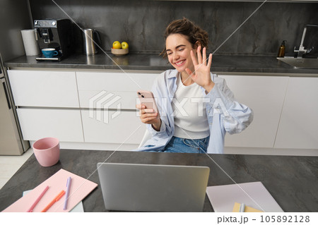 Young friendly woman, waves hand at laptop camera, video chats, holds smartphone, has conversation online 105892128