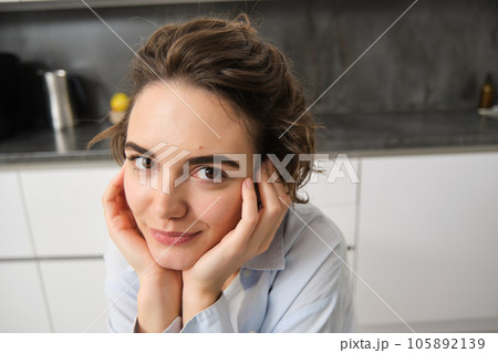 Close up portrait of young brunette woman, looks at camera and smiles, sits at home in kitchen 105892139