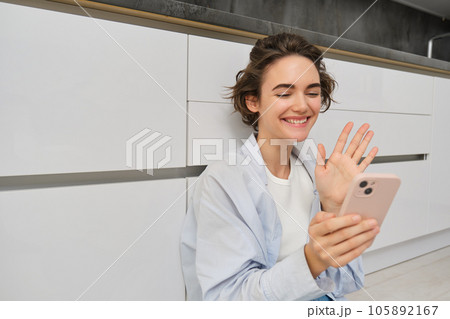 Happy girl sits on floors and video chats on smartphone app, connects to conversation on her mobile, waves hand at phone camera 105892167