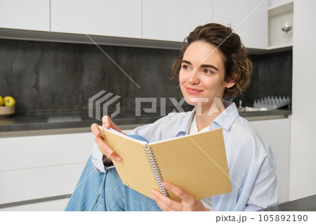 Portrait of happy smiling woman, holding notebook, studying in her kitchen, reading journal Portrait of happy smiling woman, holding notebook, studying in her kitchen, reading journal 105892190