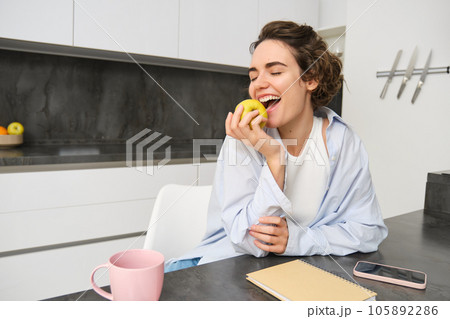 Portrait of happy brunette woman, biting an apple, eating healthy snack while sitting in kitchen Portrait of happy brunette woman, biting an apple, eating healthy snack while sitting in kitchen 105892286