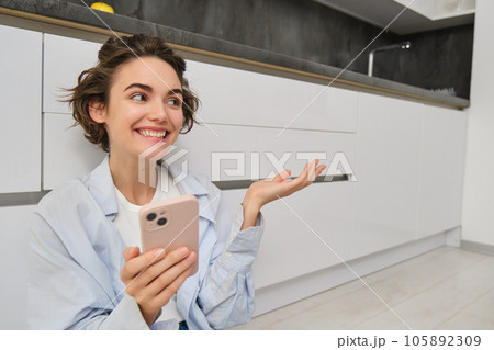 Portrait of happy young woman, shows her brand new kitchen, sits on floor, points at furniture and smiles, video chats on smartphone Portrait of happy young woman, shows her brand new kitchen, sits on floor, points at furniture and smiles, video chats on smartphone 105892309
