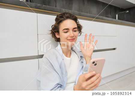 Happy girl sits on floors and video chats on smartphone app, connects to conversation on her mobile, waves hand at phone camera Happy girl sits on floors and video chats on smartphone app, connects to conversation on her mobile, waves hand at phone camera 105892314