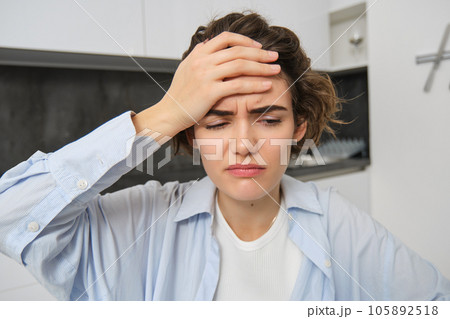 Portrait of woman with headache, sits in kitchen, touches head and grimaces, has painful migraine Portrait of woman with headache, sits in kitchen, touches head and grimaces, has painful migraine 105892518