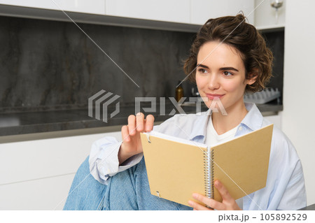 Portrait of happy smiling woman, holding notebook, studying in her kitchen, reading journal Portrait of happy smiling woman, holding notebook, studying in her kitchen, reading journal 105892529