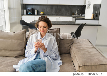 Portrait of brunette woman sits on sofa with smartphone, chats on phone, looks at her mobile app, checks messages on telephone, does online shopping from home 105892642