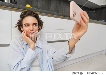 Portrait of young woman sits on kitchen floor with telephone, takes selfie on smartphone with app filters, poses for photo on mobile phone 105892657