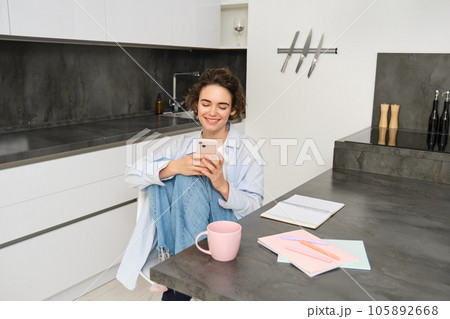 Young woman having break from work, sitting in kitchen and using smartphone, girl studies at home, sends a message on mobile phone Young woman having break from work, sitting in kitchen and using smartphone, girl studies at home, sends a message on mobile phone 105892668