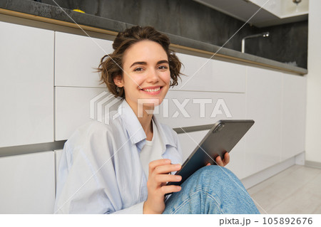 Portrait of young adult woman works on her tablet, sits with notebook on kitchen floor at home, studies in comfort 105892676
