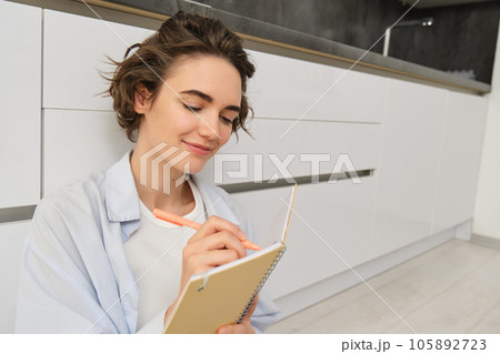 Portrait of beautiful young woman writing in journal, adding notes in planner, sitting on floor and thinking, reads her diary, smiles with pleased face expression 105892723