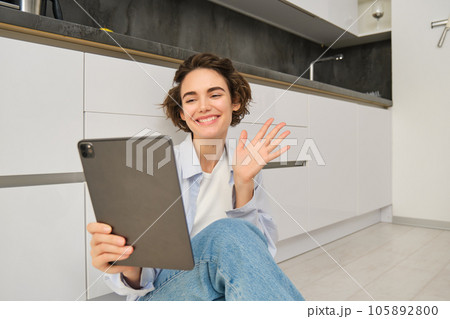 Portrait of happy young woman connects to video chat, waves at digital tablet and smiling, sits at home Portrait of happy young woman connects to video chat, waves at digital tablet and smiling, sits at home 105892800