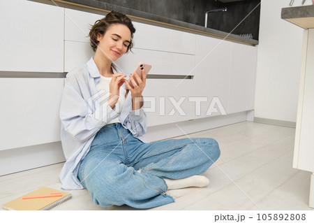 Portrait of cute smiling woman, sitting on floor at home with crossed legs, looks at mobile phone, chats on smartphone app, watches videos in application Portrait of cute smiling woman, sitting on floor at home with crossed legs, looks at mobile phone, chats on smartphone app, watches videos in application 105892808