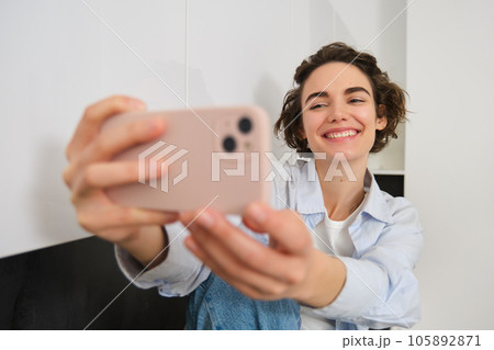 Portrait of beautiful brunette girl, takes selfie on smartphone in her kitchen, posing for photo on mobile phone 105892871