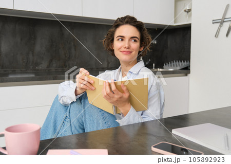 Young woman sitting in kitchen, reading her notes in notebook. Smiling girl at home, doing homework, studying 105892923