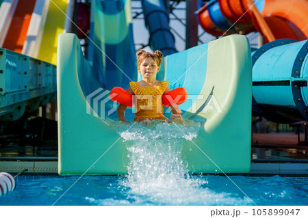 Little preschool girl on water slide in aqua park. Happy child having fun on water slides on family summer vacation in tropical resort. Amusement park with wet playground for young child and baby. Little preschool girl on water slide in aqua park. Happy child having fun on water slides on family summer vacation in tropical resort. Amusement park with wet playground for young child and baby. 105899047