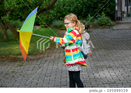 Little girl on way to elementary school or kindergarden. Preschool Child with colorful rainbow umbrella and waterproof jacket with school bag. Kid walking in autumn shower. Outdoor fun by any weather Little girl on way to elementary school or kindergarden. Preschool Child with colorful rainbow umbrella and waterproof jacket with school bag. Kid walking in autumn shower. Outdoor fun by any weather 105899159